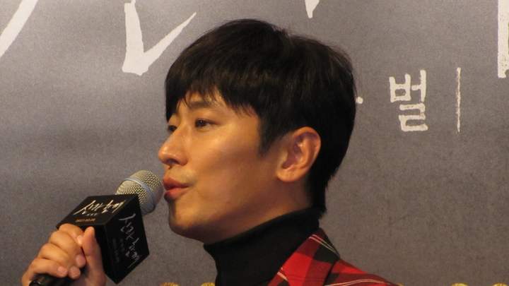 Ju Ji-hoon de paletó xadrez vermelho e gola alta preta, falando ao microfone