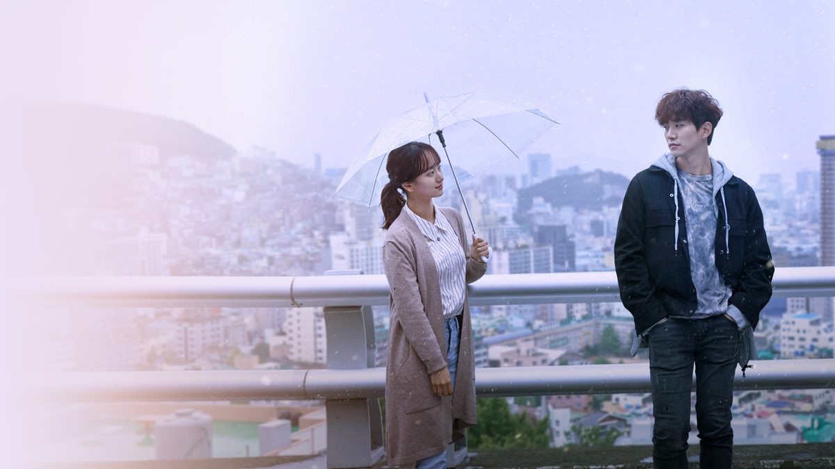 Uma jovem de cardigã bege segura um guarda-chuva transparente e olha para um rapaz de jaqueta preta e moletom cinza, os dois junto a uma grade num mirante com a cidade ao fundo sob chuva fina