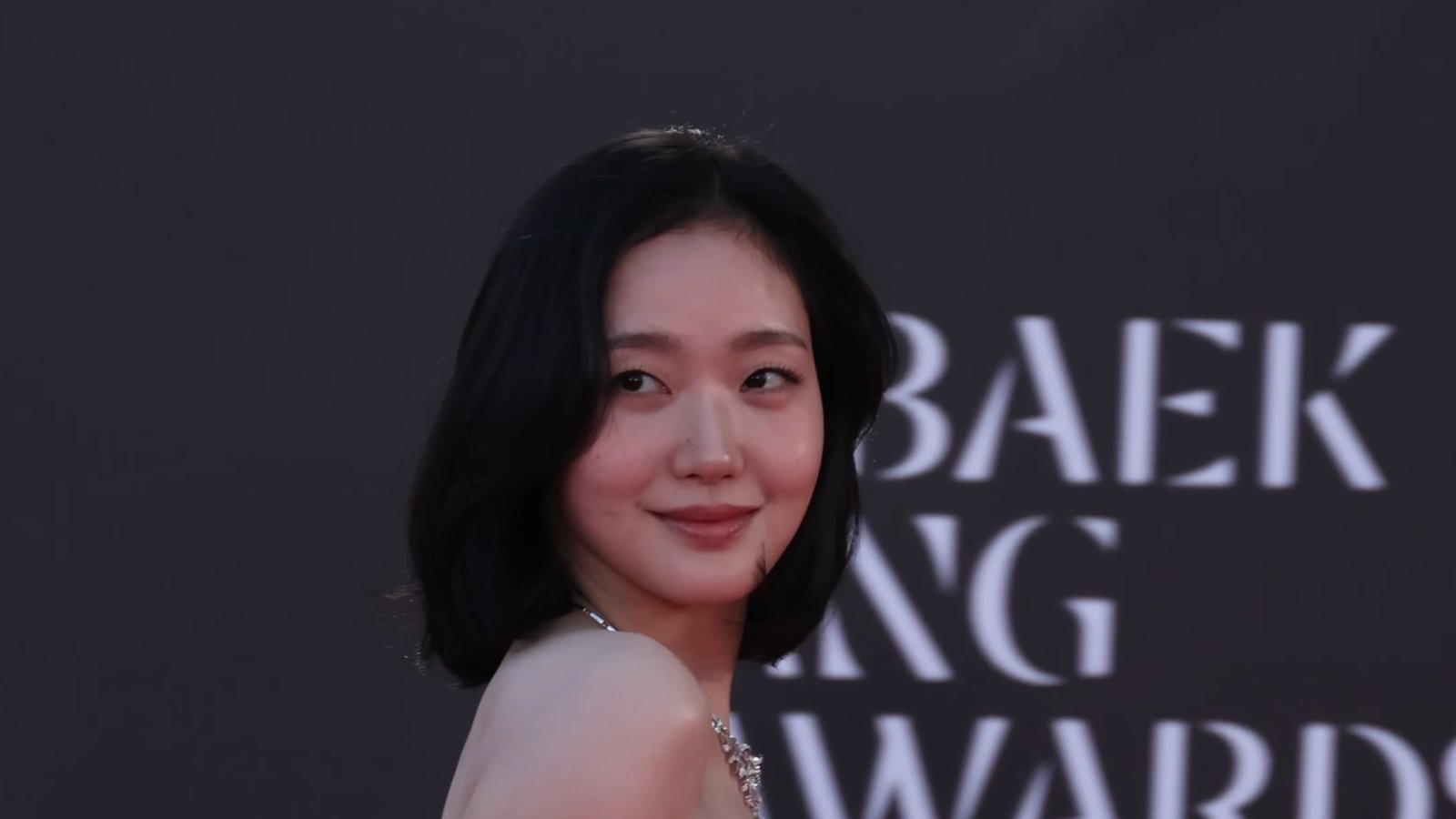Kim Go-eun de cabelo curto e vestido branco tomara que caia sorri diante de painel do Baeksang