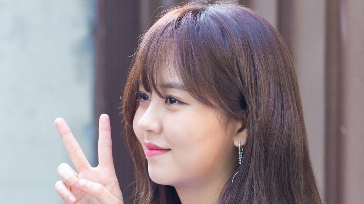 Kim So-hyun de perfil, cabelo castanho longo e camiseta branca, sorri e faz sinal de paz com a mão