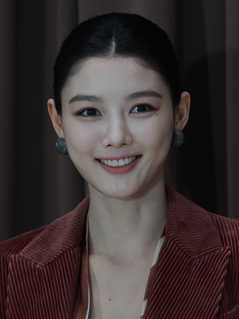 Kim Yoo-jung de cabelo preso e blazer de veludo vinho sorri diante de cortina escura