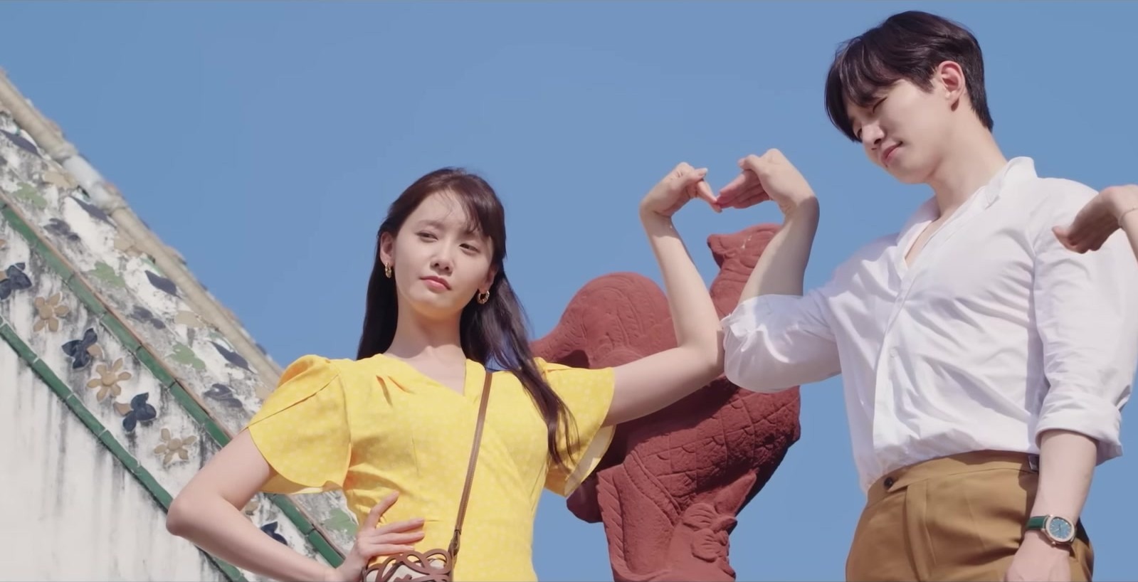 Im Yoon-ah, de blusa amarela, e Lee Jun-ho, de camisa branca, fazem um coração com os braços diante de um céu azul, em cena do trailer de King the Land