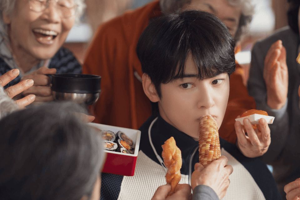 Rapaz de cabelo preto e franja, de blusa branca, morde uma espiga de milho enquanto senhoras sorridentes em volta oferecem kimbap, batata-doce e outros petiscos