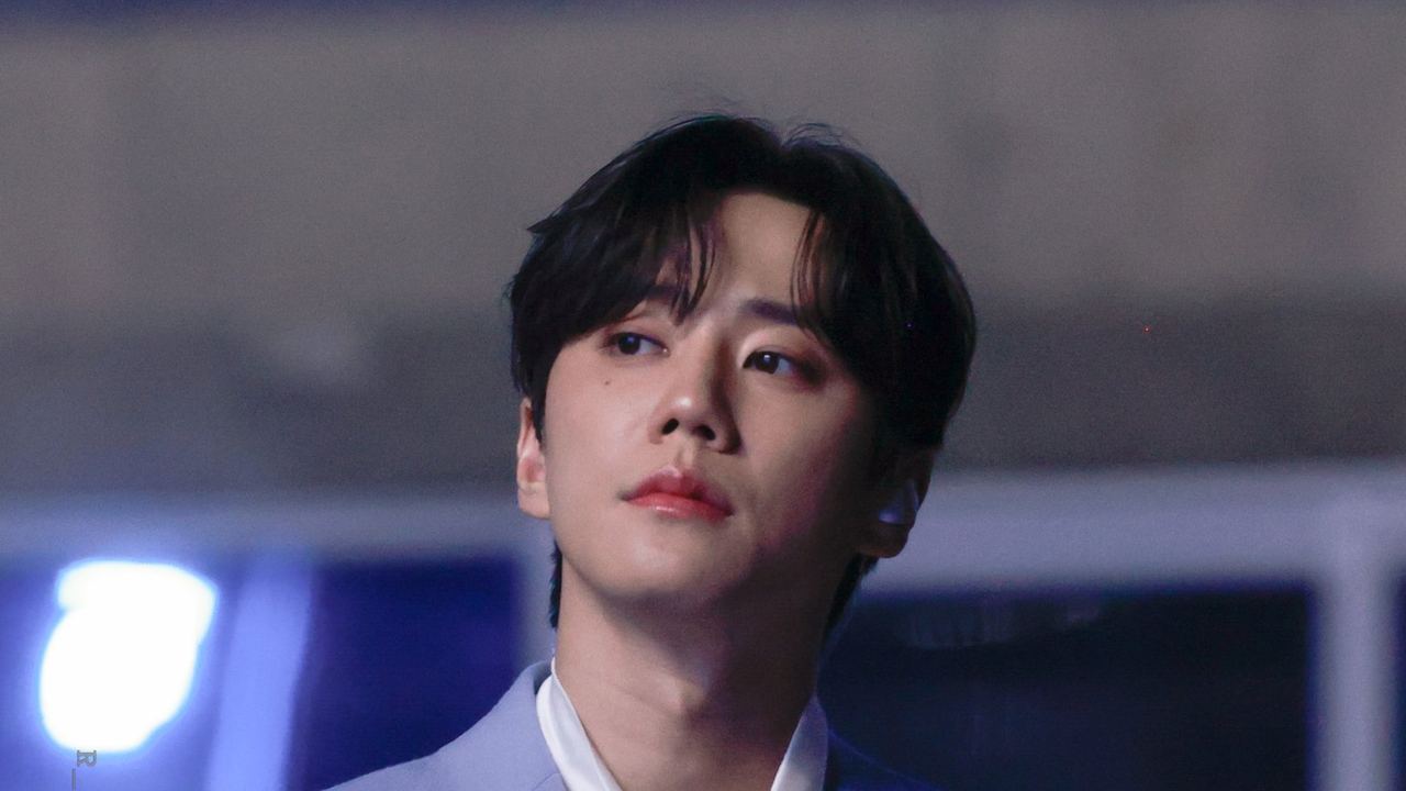 Lee Jun-young de terno azul-claro e camisa branca olha para o lado num palco com luzes azuis ao fundo
