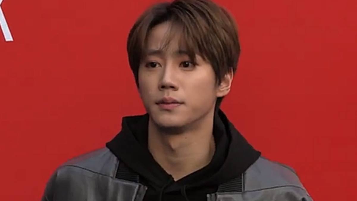 Lee Jun-young de jaqueta cinza e moletom preto de capuz posa diante de uma parede vermelha