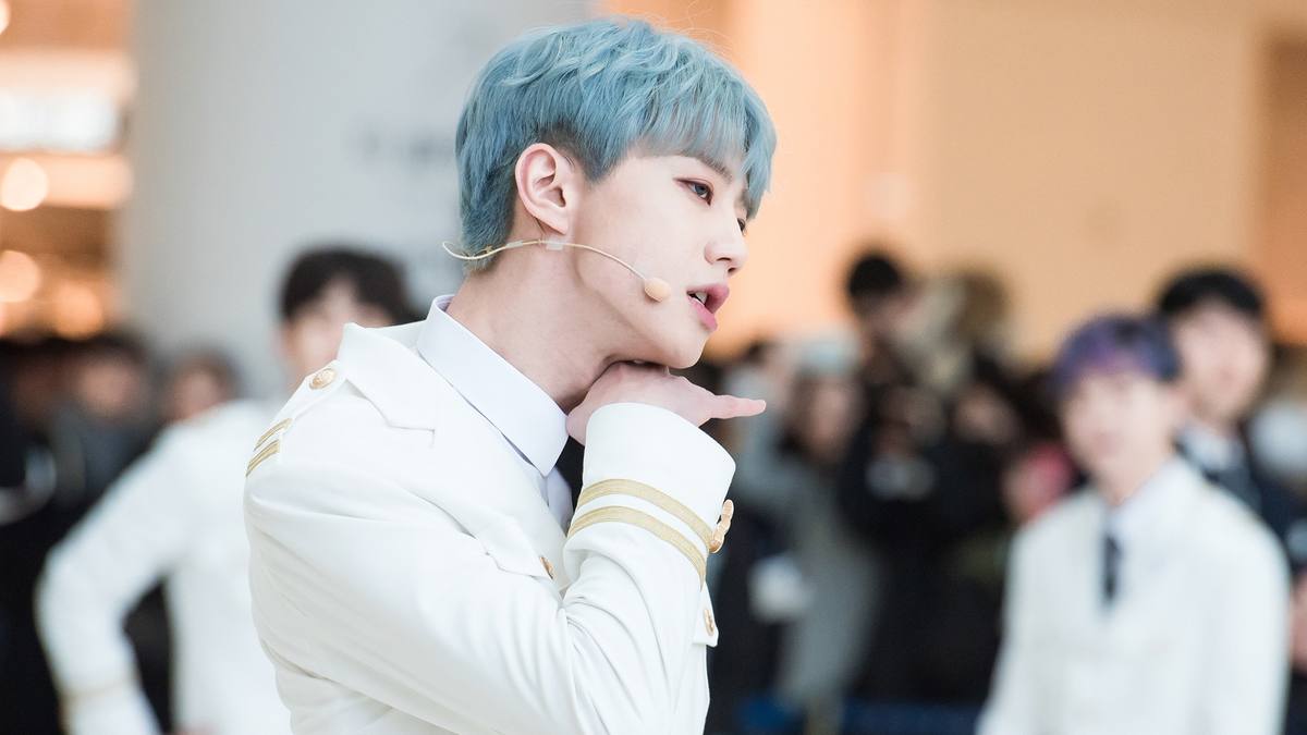 Lee Jun-young de cabelo azul e roupa branca de detalhes dourados faz pose com a mão no queixo, com outros rapazes ao fundo