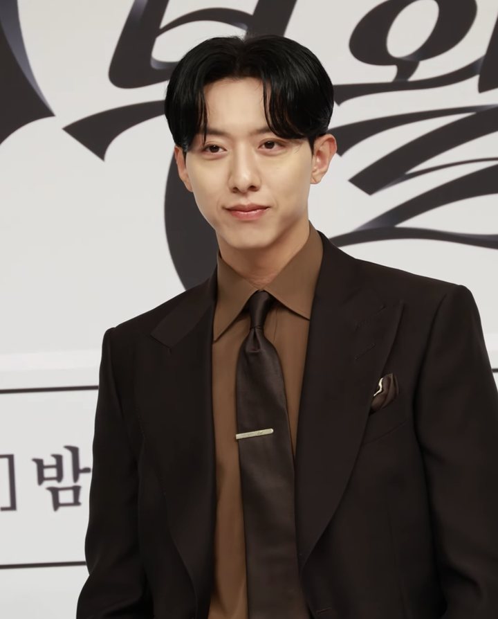 Homem de cabelo preto com franja repartida ao meio, camisa marrom, gravata marrom brilhante com prendedor dourado e terno marrom-escuro, sorrindo de leve diante de painel branco com letras pretas