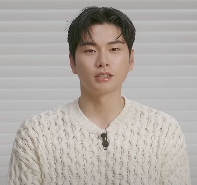 Lee Yi-kyung de suéter branco de tricô, falando para a câmera diante de uma parede clara