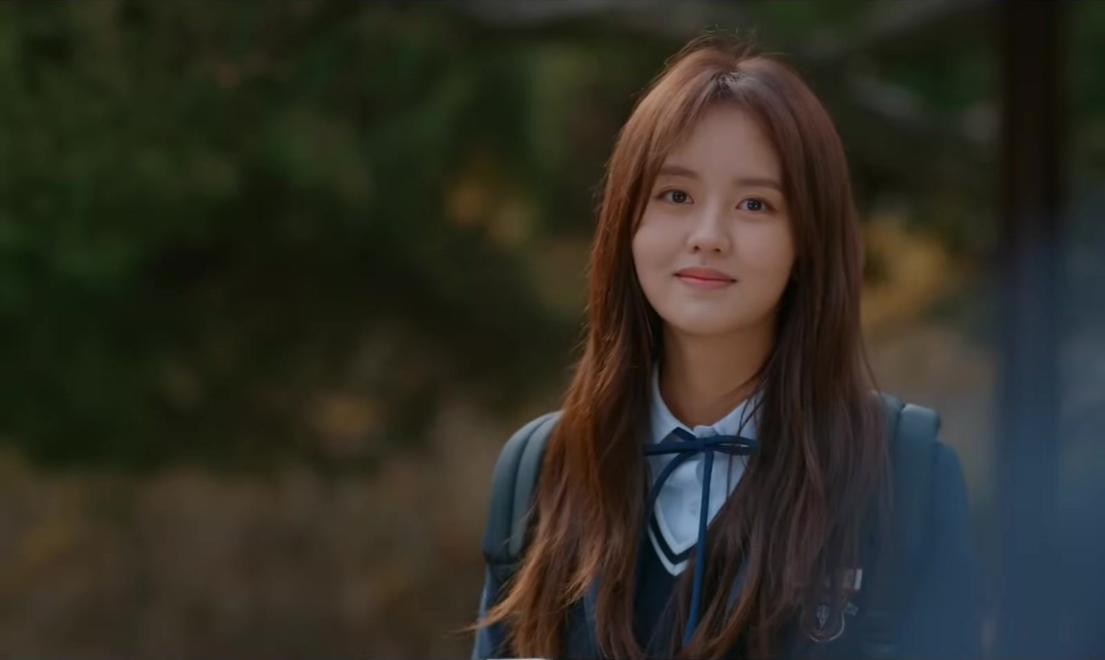 Kim So-hyun, de uniforme azul-marinho com laço, sorri de leve num parque, em quadro do trailer de Love Alarm