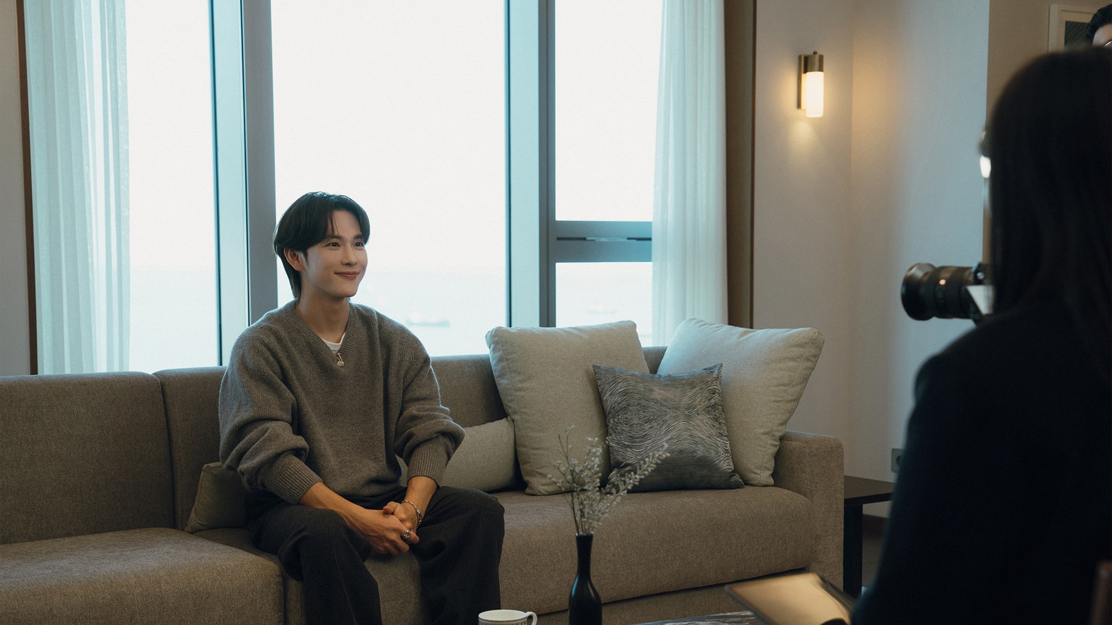 Yim Si Wan sorrindo sentado num sofá, de frente para uma câmera, em cena de entrevista de Love in Disguise