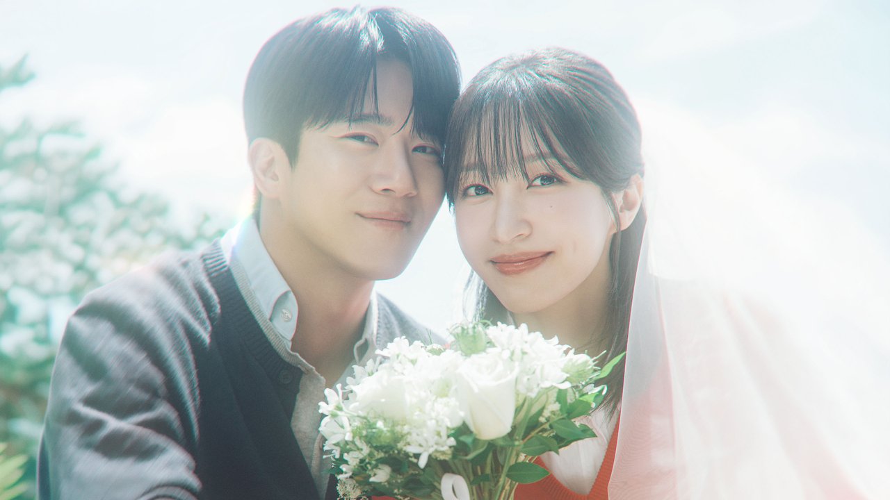 Ha Seok-jin, de cardigã cinza escuro, e Ahn Hee-yeon, de véu transparente e buquê de flores brancas, encostam as cabeças e sorriem para a câmera sob luz clara