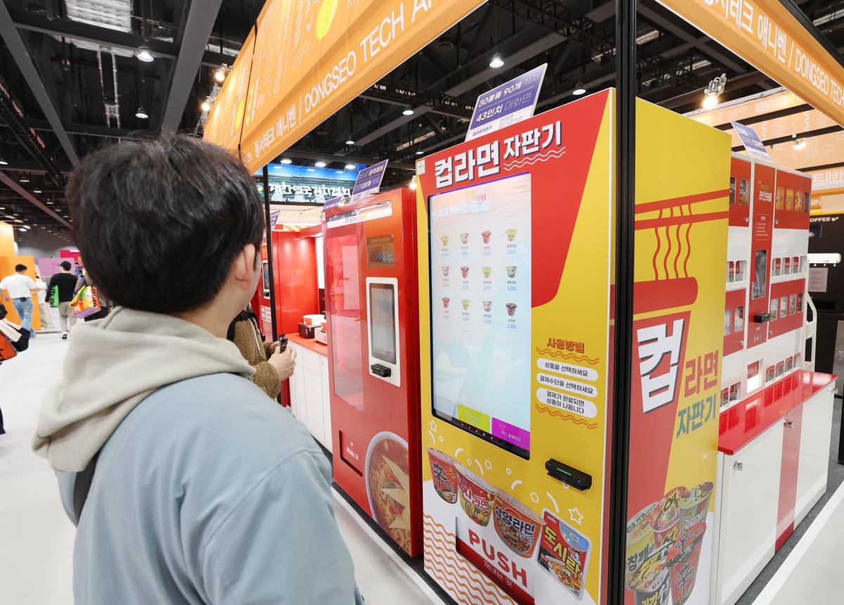 Visitante de costas olha uma máquina de venda de ramyeon em copo, de painel amarelo e vermelho, num corredor da feira