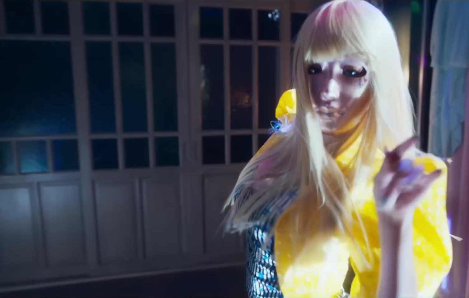 Figura de peruca loira longa, máscara no rosto e roupa amarela, apontando, em corredor azulado, em quadro do trailer oficial de Mask Girl