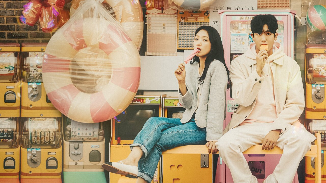Park Shin-hye de cardigã cinza e Park Hyung-sik de roupa clara sentados sobre máquinas de brinquedo, cada um com um picolé, diante de uma parede cheia de máquinas de cápsulas e uma boia listrada