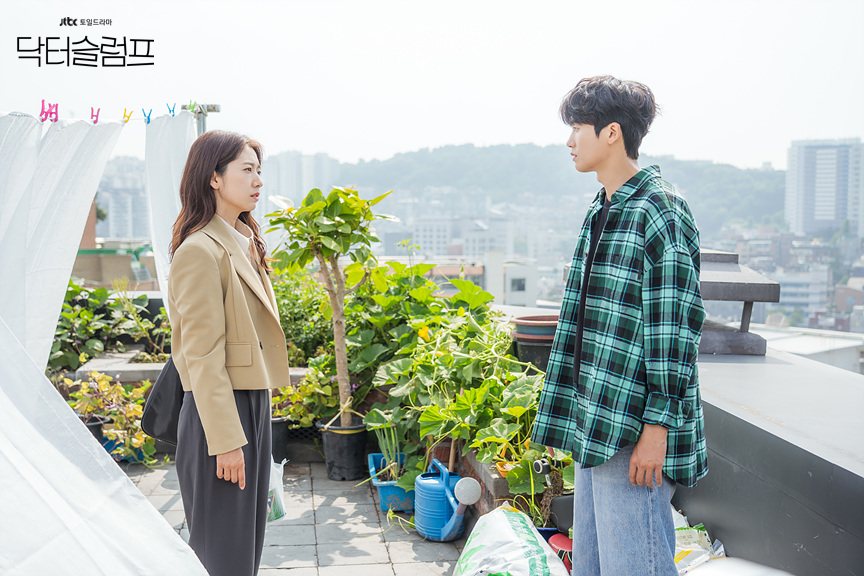 Park Shin-hye de blazer bege e Park Hyung-sik de camisa xadrez verde frente a frente num terraço com plantas em vasos e vista de prédios