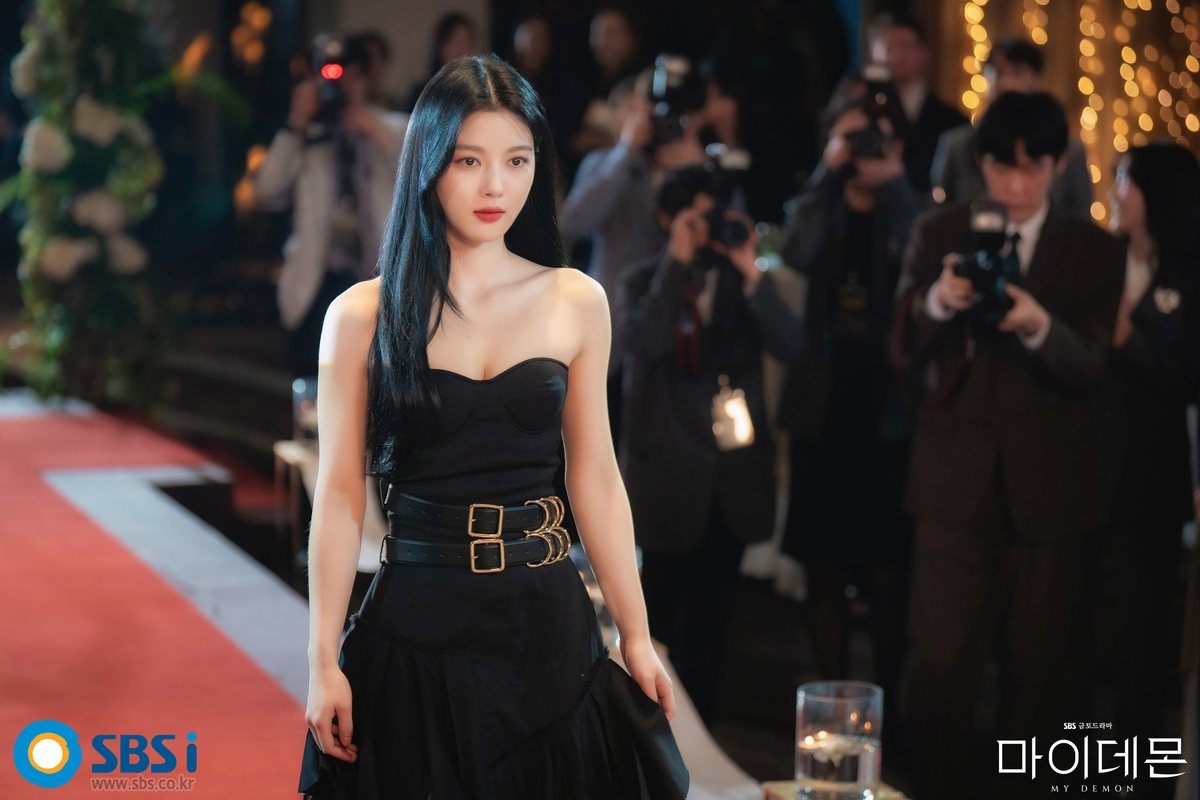 Kim You-jung como Do-hee, de vestido preto tomara que caia com dois cintos, caminha por um tapete vermelho diante de fotógrafos
