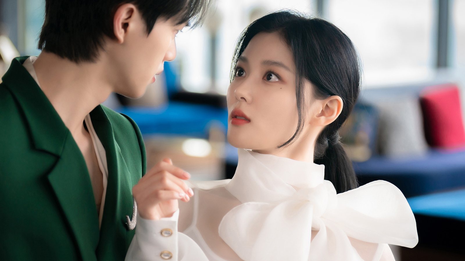 Song Kang, de paletó verde, e Kim You-jung, de blusa branca com laço, se encaram de perto num salão com sofás azuis