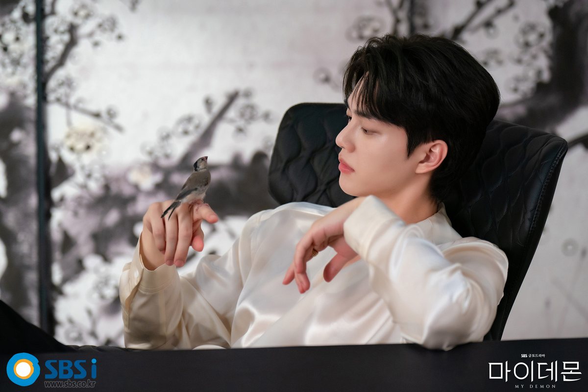 Song Kang como Gu-won, de camisa de cetim creme, sentado numa poltrona preta observa um pássaro pousado em sua mão, diante de um painel com galhos floridos