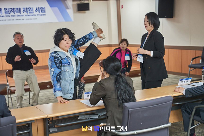 Lee Jung-eun de jaqueta jeans estampada erguendo a perna bem alto numa sala de seleção para estágio sênior, diante de avaliadores e outros candidatos idosos