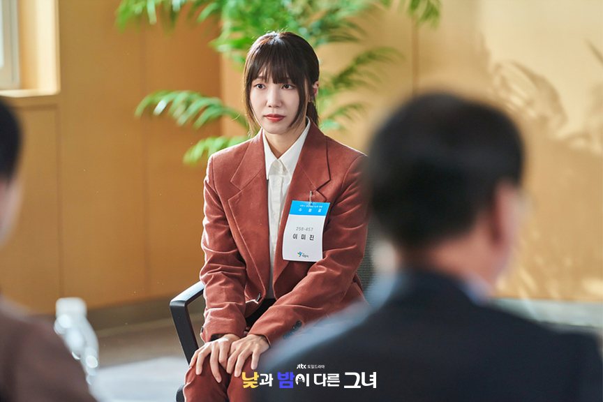 Jeong Eun-ji como Lee Mi-jin, de terninho cor de ferrugem e crachá de candidata, sentada diante de entrevistadores desfocados numa sala com plantas