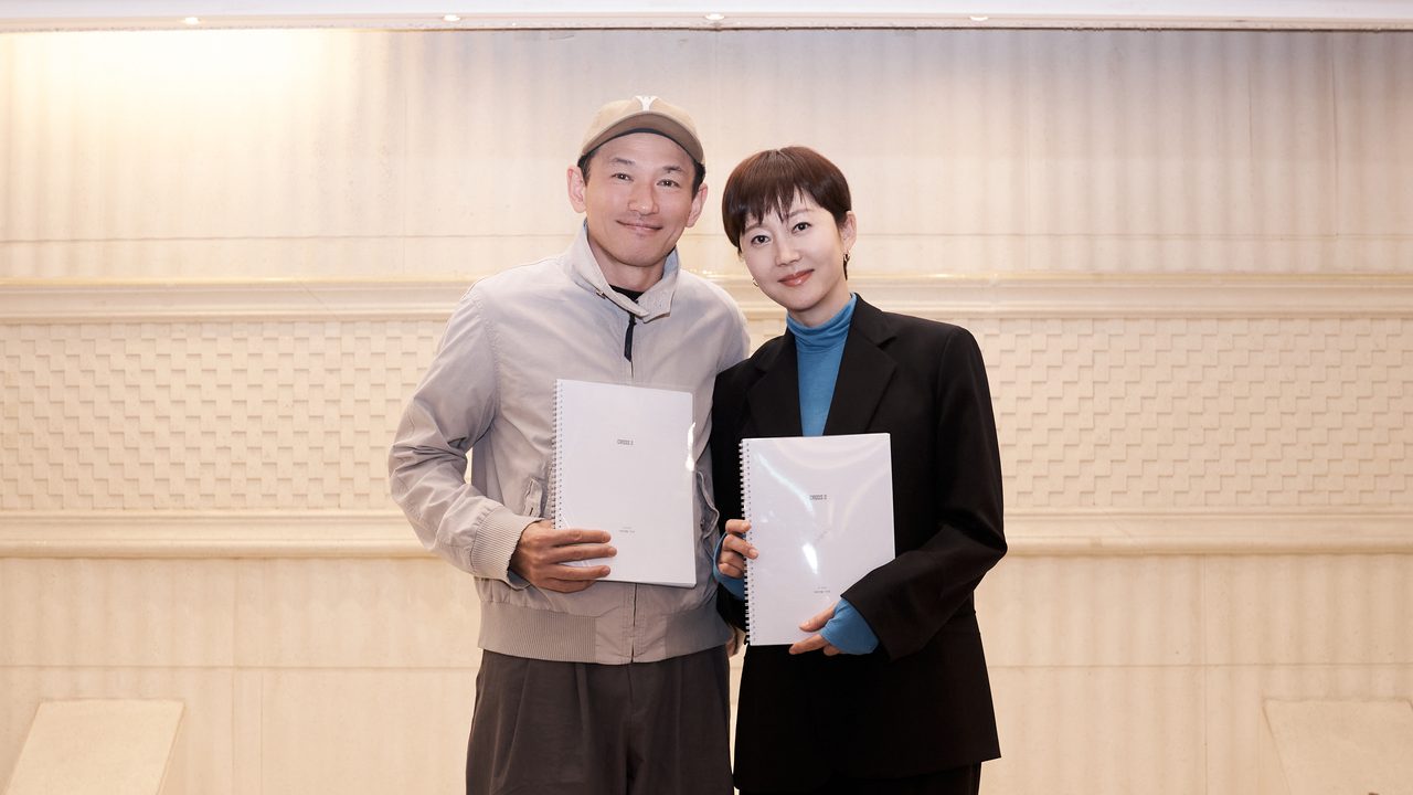 Hwang Jung-min de boné e jaqueta cinza ao lado de Yum Jung-ah de blazer preto, os dois segurando o roteiro