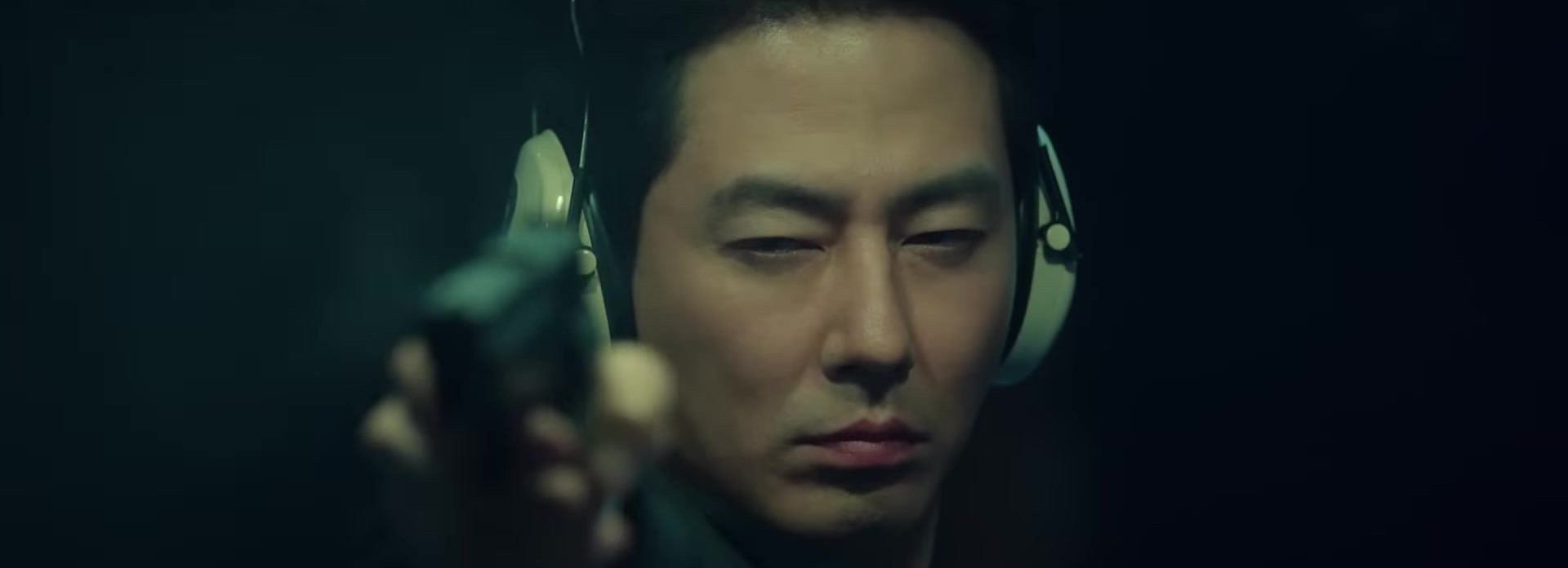 Close de um homem de fones de proteção auditiva verdes, com uma arma desfocada em primeiro plano, em quadro do trailer oficial de Moving, do Disney+