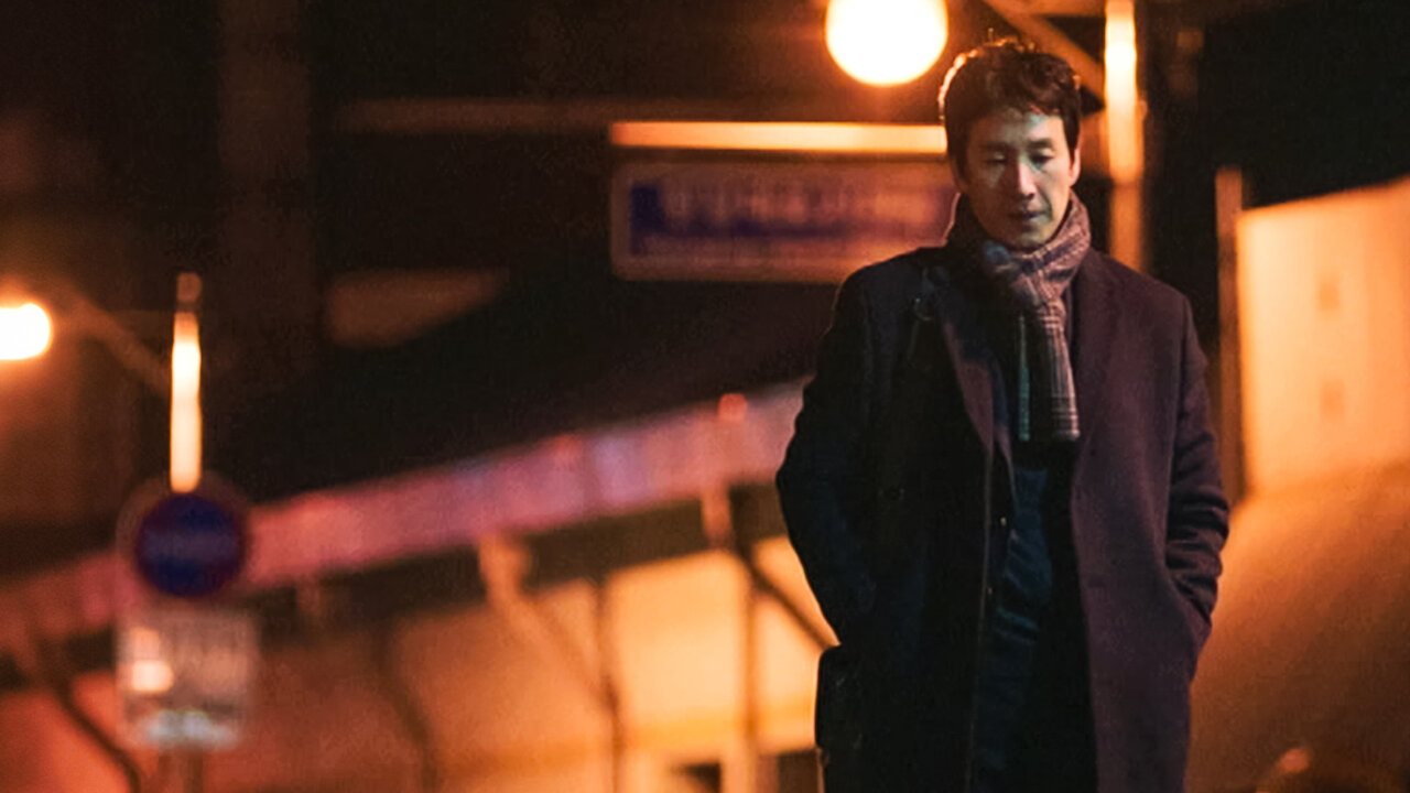 Arte de divulgação de My Mister na Netflix: homem de sobretudo escuro e cachecol xadrez andando à noite, de cabeça baixa e mãos nos bolsos, com luzes alaranjadas desfocadas e uma placa de rua ao fundo
