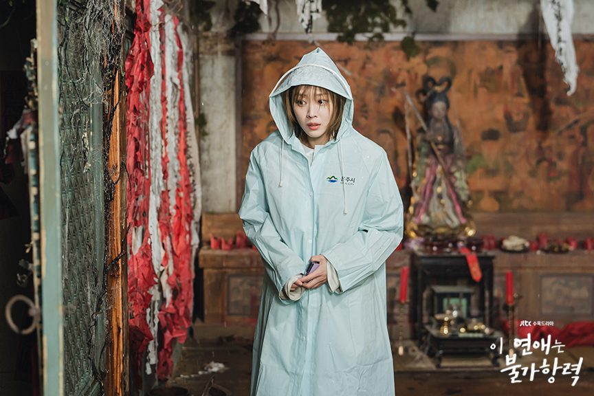 Cho Bo-ah como Lee Hong-jo, de capa de chuva azul-clara com o símbolo da prefeitura, dentro de um santuário com tiras de papel vermelho e branco, velas e uma estátua no altar