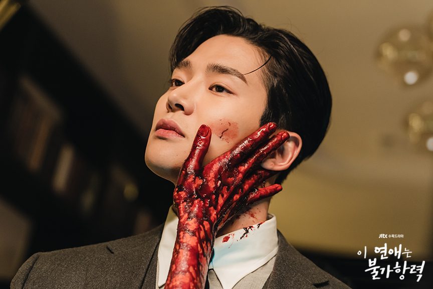 Rowoon como Jang Sin-yu, de terno cinza, com uma mão vermelha, de aspecto ensanguentado, segurando o lado esquerdo do rosto dele