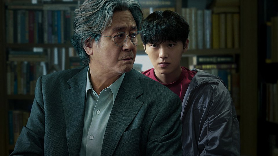Choi Min-sik de cabelo grisalho, óculos e paletó cinza olha de lado numa biblioteca, com Choi Hyun-wook de moletom vermelho e jaqueta escura logo atrás dele