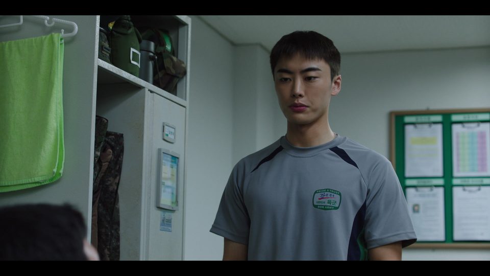 Jovem de camiseta cinza do exército de pé num alojamento, ao lado de armários com toalha verde pendurada