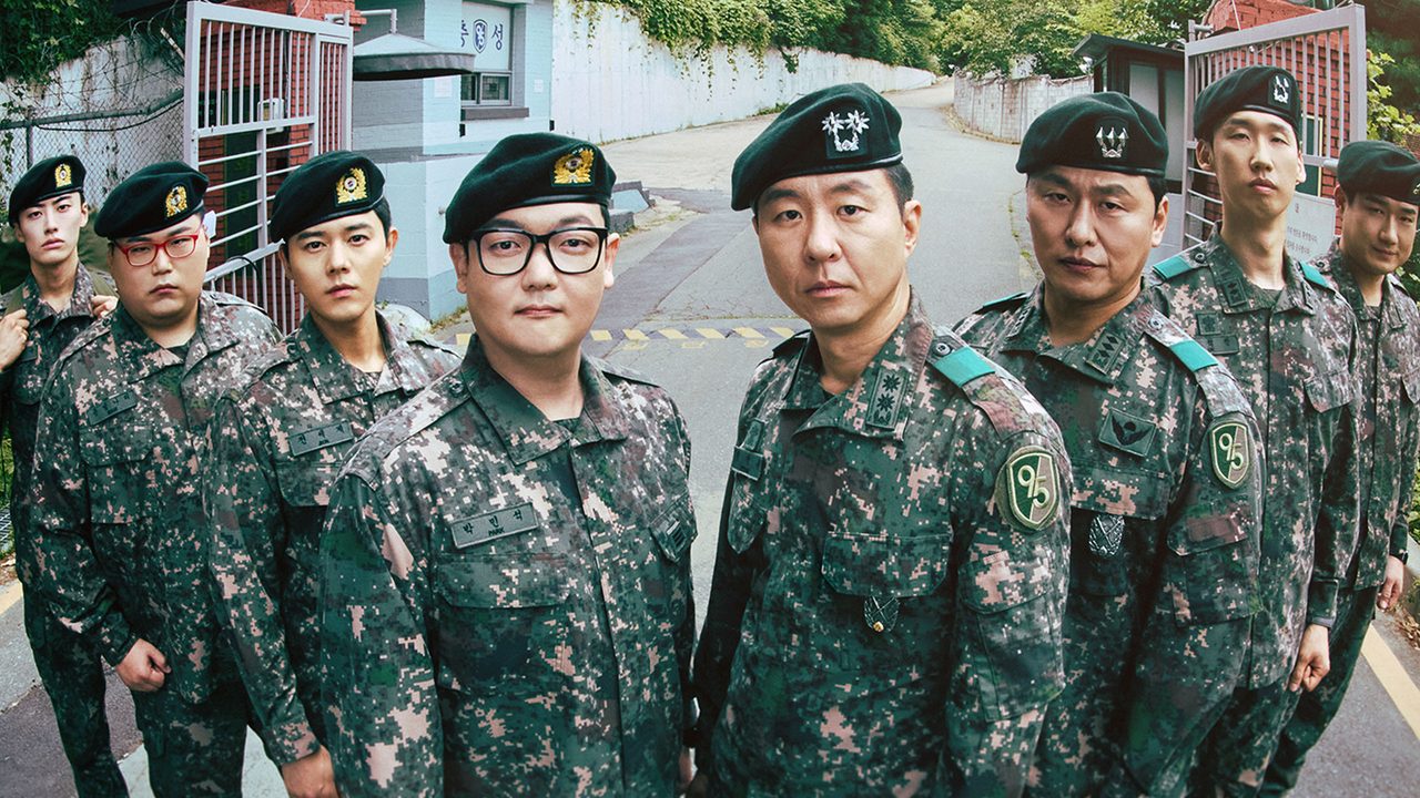 Oito soldados de uniforme camuflado e boina preta encaram a câmera diante do portão aberto de um quartel, numa rua em subida cercada de muros e árvores; no centro, um homem de óculos de armação preta