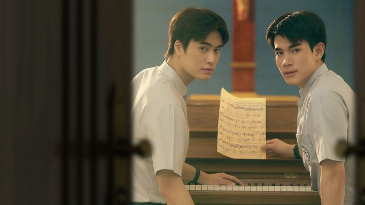 Dois rapazes de uniforme escolar branco junto a um piano, um deles segurando uma partitura, vistos por uma porta entreaberta