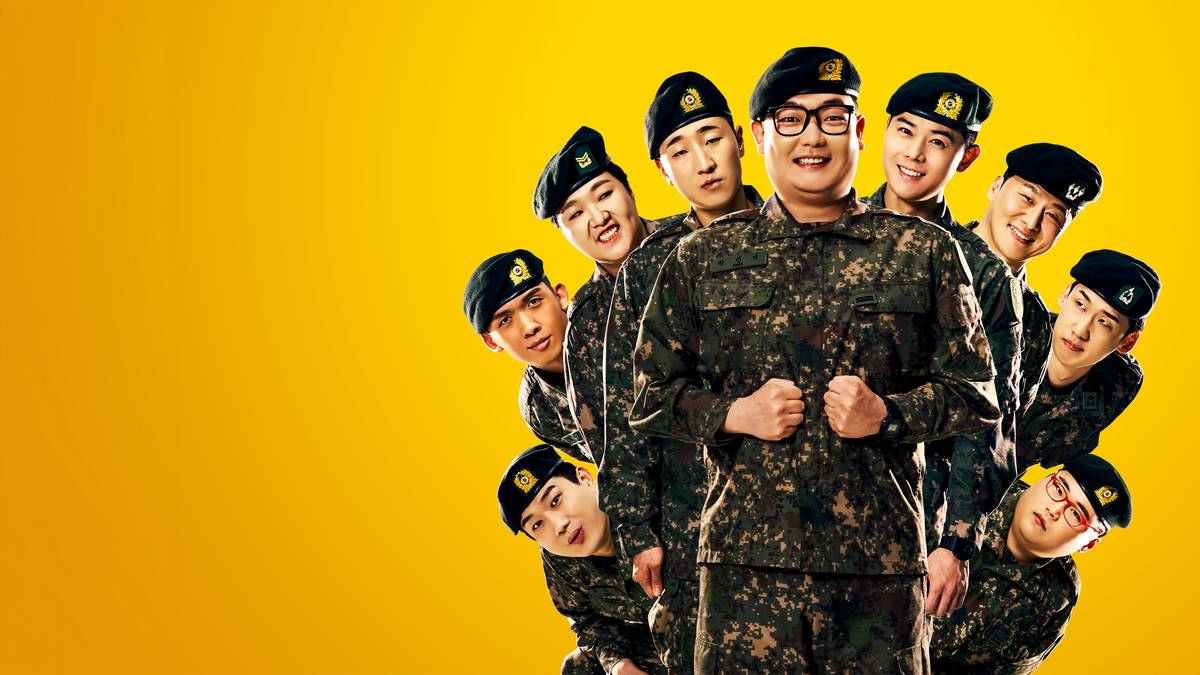 Grupo de soldados de farda camuflada e boina preta em torno de um soldado de óculos que sorri com os punhos fechados no peito, sobre fundo amarelo