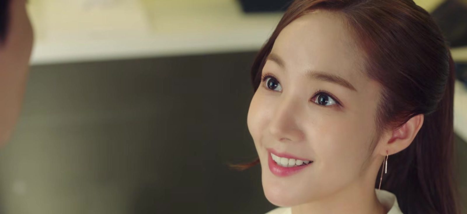Park Min Young de perfil, sorrindo, com cabelo preso e brinco longo, em quadro do trailer oficial de O Que Há de Errado com a Secretaria Kim