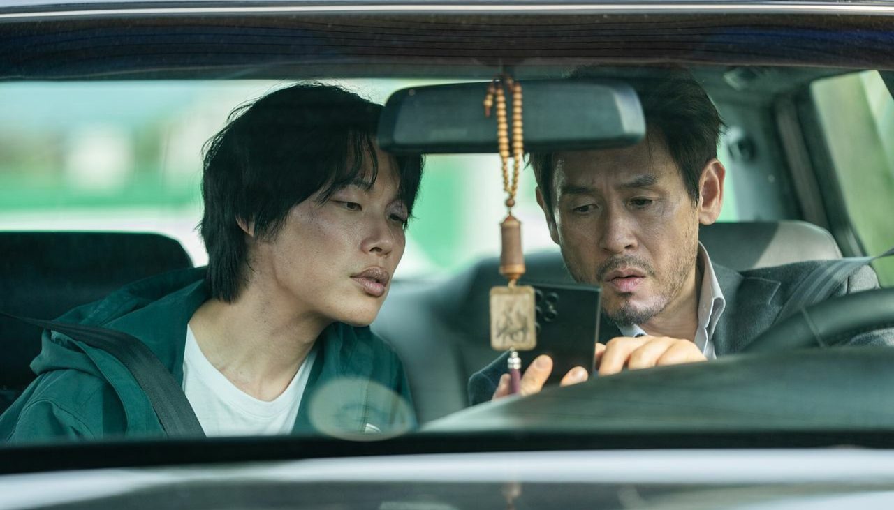Ryu Jun-yeol, de jaqueta verde, e Sul Kyung-gu, de terno, olham juntos para um celular dentro de um carro, vistos pelo para-brisa