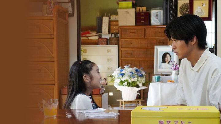 Menina de cabelo comprido e homem de camisa branca sentados à mesa de uma sala, diante de um retrato de mulher com flores brancas e azuis