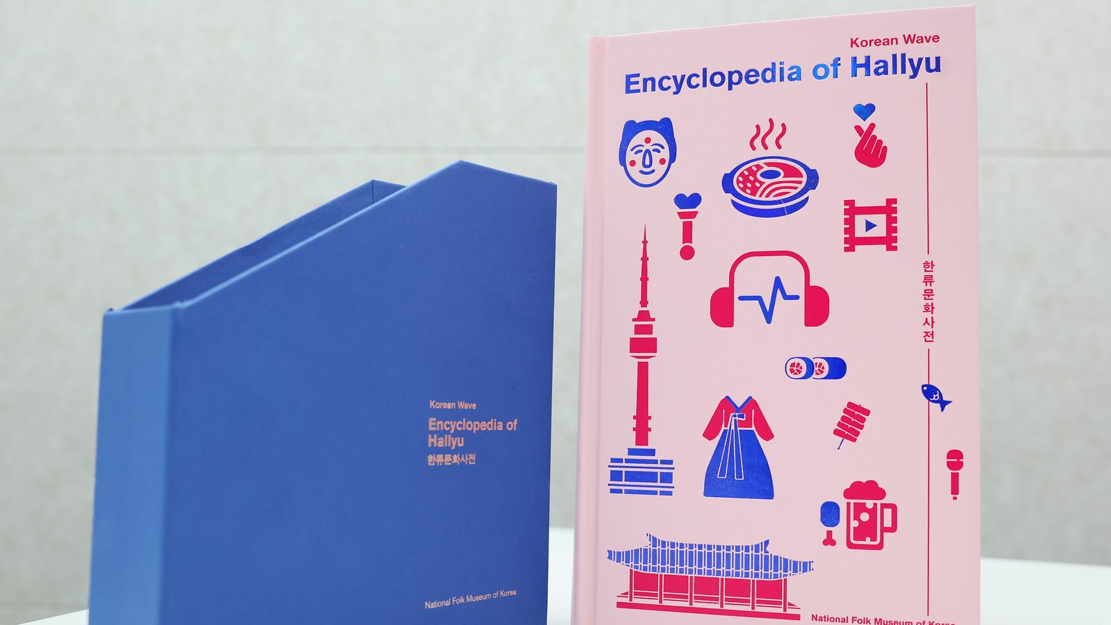 Livro de capa rosa com ícones coreanos e o título Encyclopedia of Hallyu ao lado de uma caixa azul, sobre mesa branca