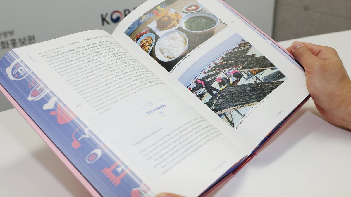 Mãos segurando a Encyclopedia of Hallyu aberta, com texto à esquerda e fotos de comida coreana e de um barco à direita