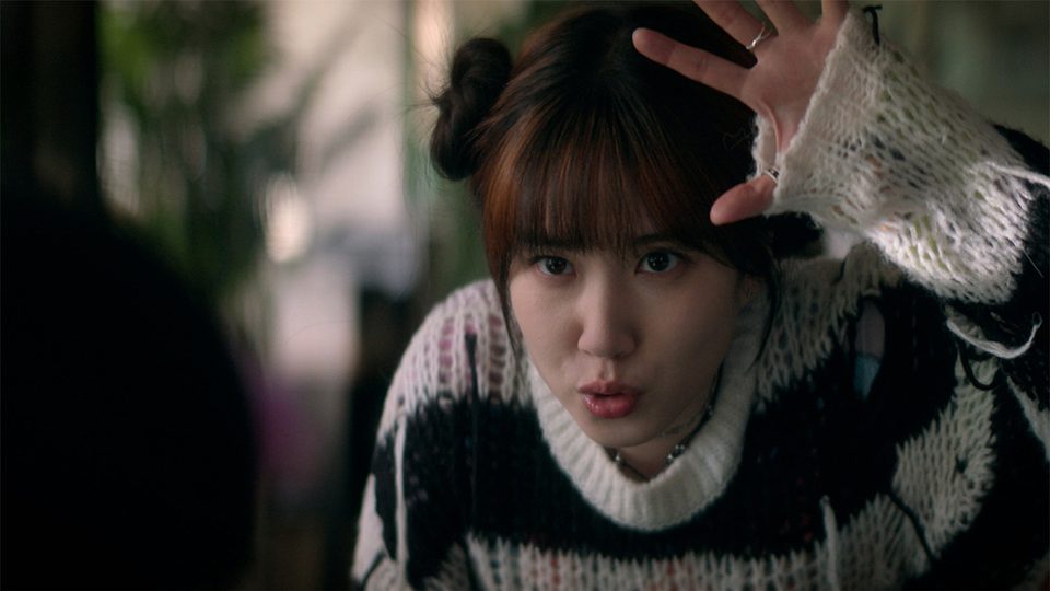 Park Eun-bin de suéter listrado preto e branco, coque no cabelo, ergue a mão aberta acima da testa com os lábios em bico