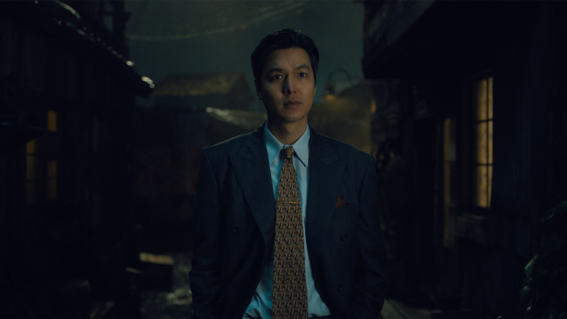 Lee Min Ho, de terno azul e gravata estampada, parado numa viela escura em cena da 2ª temporada de Pachinko, do Apple TV+