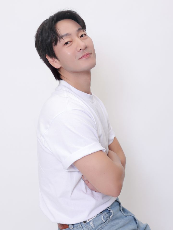 Park Hae-soo de camiseta branca e calça jeans, sentado de braços cruzados num estúdio claro