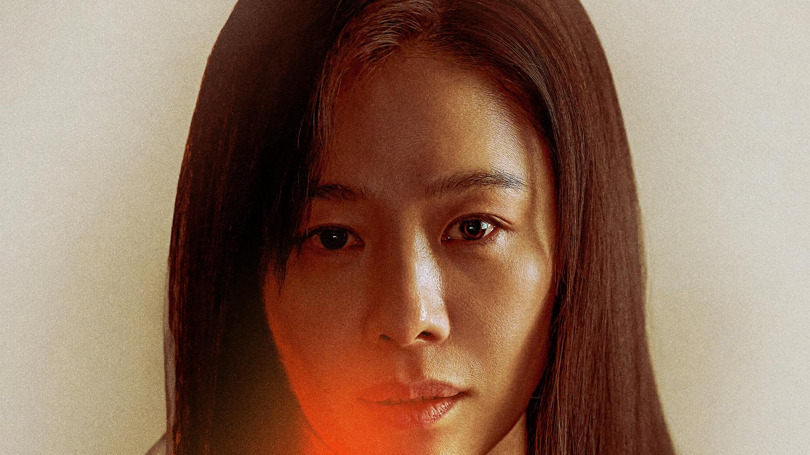 Kim Hyun-joo em close, de cabelo liso e escuro e olhar fixo na câmera, diante de fundo bege com luz vermelha sob o queixo
