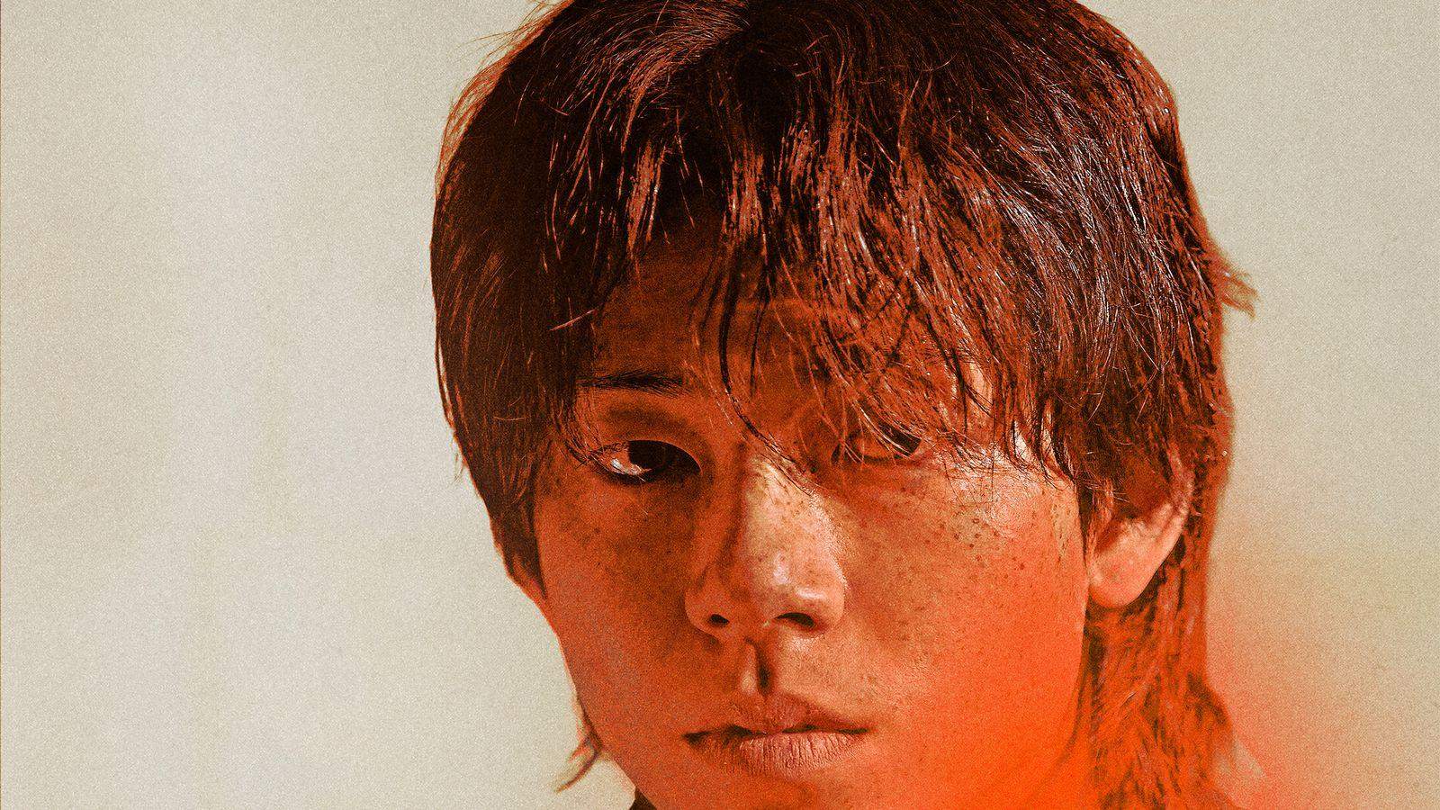 Bae Hyeon-seong em close, de cabelo despenteado e rosto cansado, olhando de lado diante de fundo bege com luz vermelha