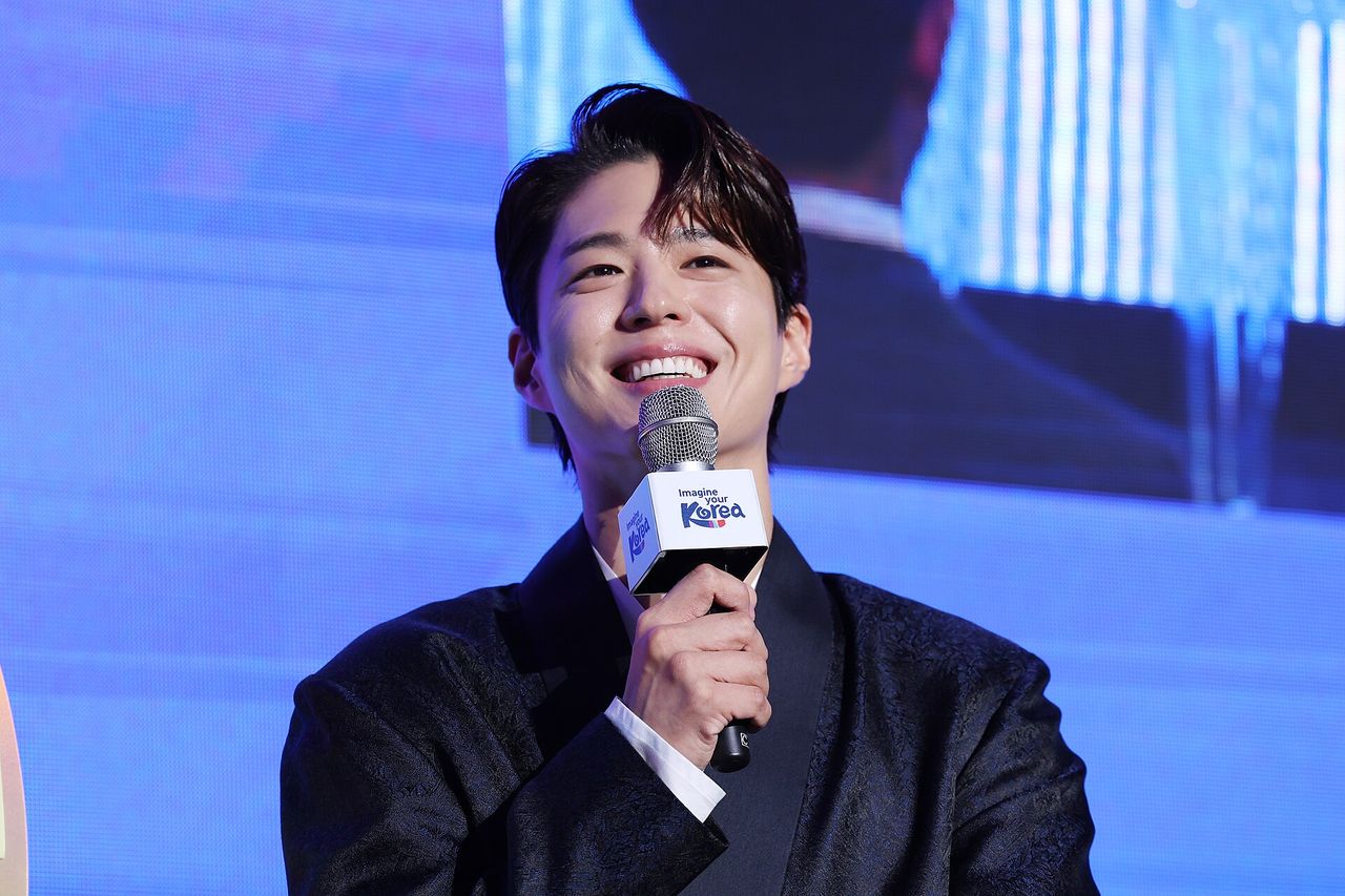 Homem de cabelo castanho e paletó escuro sorrindo e segurando um microfone com a marca Never Ending Korea, diante de um telão azul
