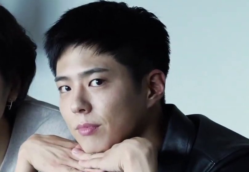 Park Bo Gum de jaqueta de couro preta apoia o queixo nas mãos e olha para a câmera diante de fundo cinza-azulado