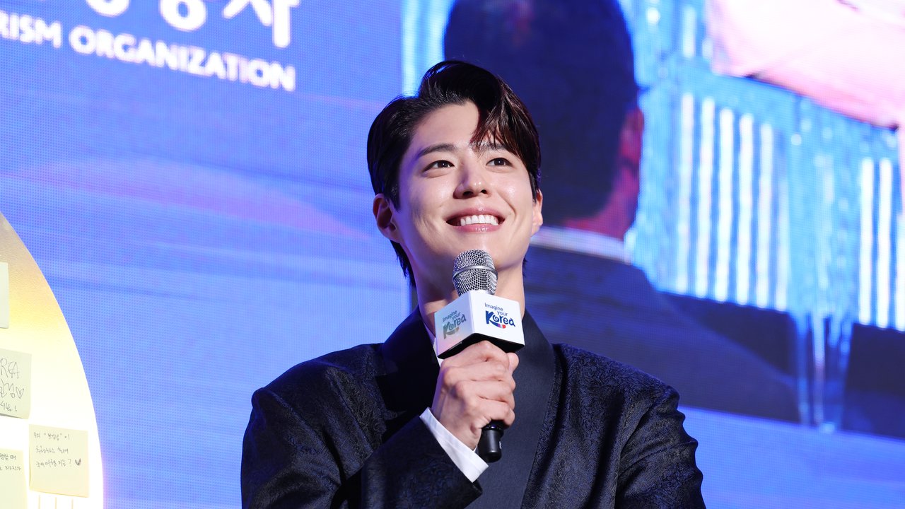 Park Bo Gum sorri de terno escuro e segura um microfone com a marca Imagine your Korea diante de um telão azul