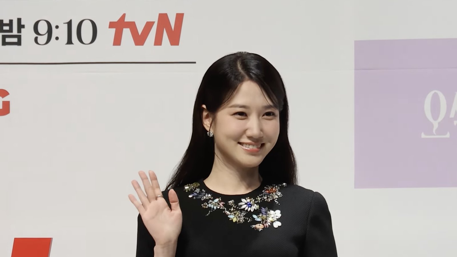 Park Eun-bin de vestido preto acena diante de painel branco e lilás com logotipos da tvN