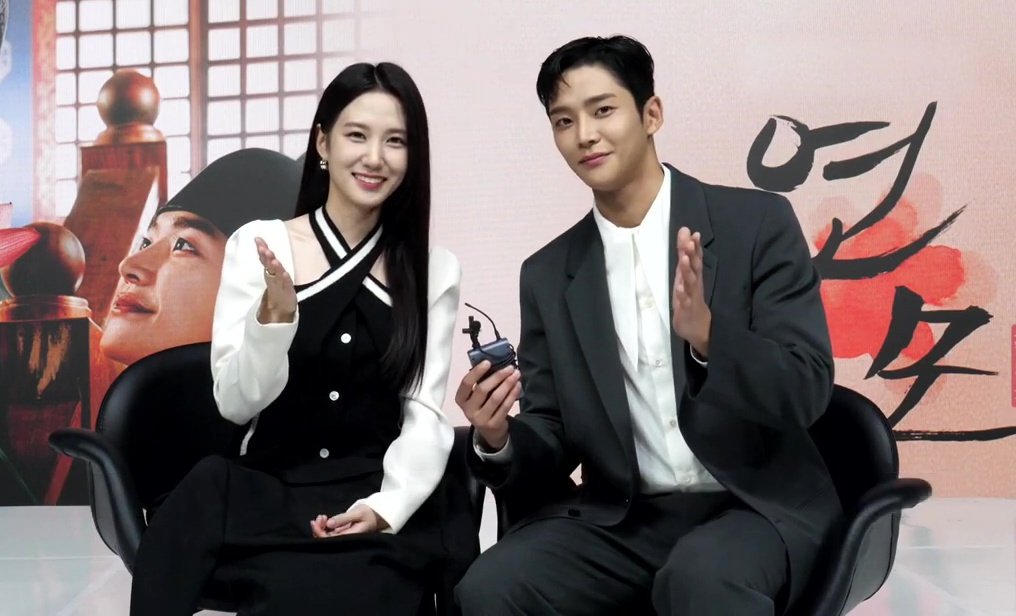 Park Eun-bin e Rowoon sentados lado a lado acenam diante de cartaz de O Rei de Porcelana