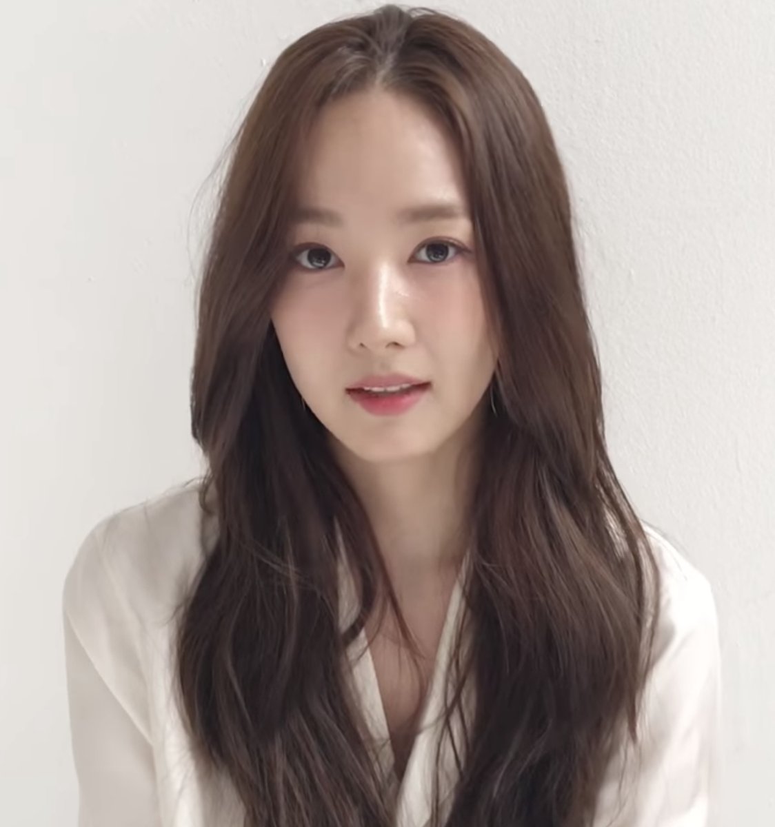 Park Min-young de cabelo longo ondulado e blusa branca diante de parede branca